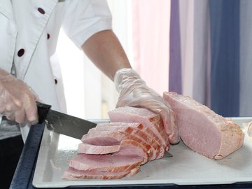 Das Foto zeigt einen Mann in Kochkleidung, der einen Schinken in Scheiben schneidet