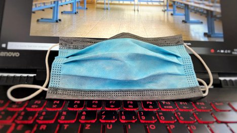 Auf dem Foto ist ein Mund-Nasen-Schutz abgebildet, der auf einem Laptop liegt, auf dessen Bildschirm ein Klassenraum zu sehen ist. 