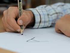 DIE LINKE_PM_100 Jahre Grundschule Das Bildelement ist eine Fotografie, die in einem Ausschnitt die Hände eines Jungen, der ein helles kariertes, langärmeliges Hemd trägt, sowie ein Blatt Papier zeigt. Mit einem schwarz-gelben Bleistift schreibt der Junge den Buchstaben A. Das große A hat er bereits vollständig geschrieben, das kleine a schreibt er gerade.