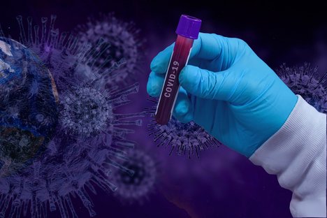 Das Foto zeigt im Hintergrund Illustrationen des Corona-Virus' und im Vordergrund eine Hand, die ein Test-Rohr hält. 