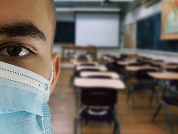 Das Foto zeigt einen Jungen mit einer Mund-Nase-Maske, im Hintergrund ist ein Klassenzimmer zu sehen. 