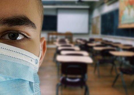 Das Foto zeigt einen Jungen mit einer Mund-Nase-Maske, im Hintergrund ist ein Klassenzimmer zu sehen. 