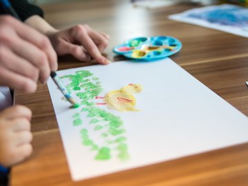 Das Bild zeigt eine erwachsene Person, die einem Kind beim Malen hilft. 