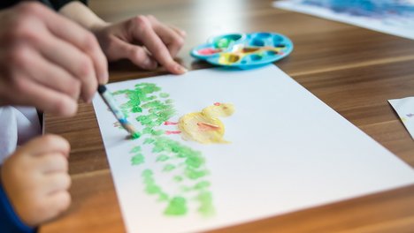 Das Bild zeigt eine erwachsene Person, die einem Kind beim Malen hilft. 